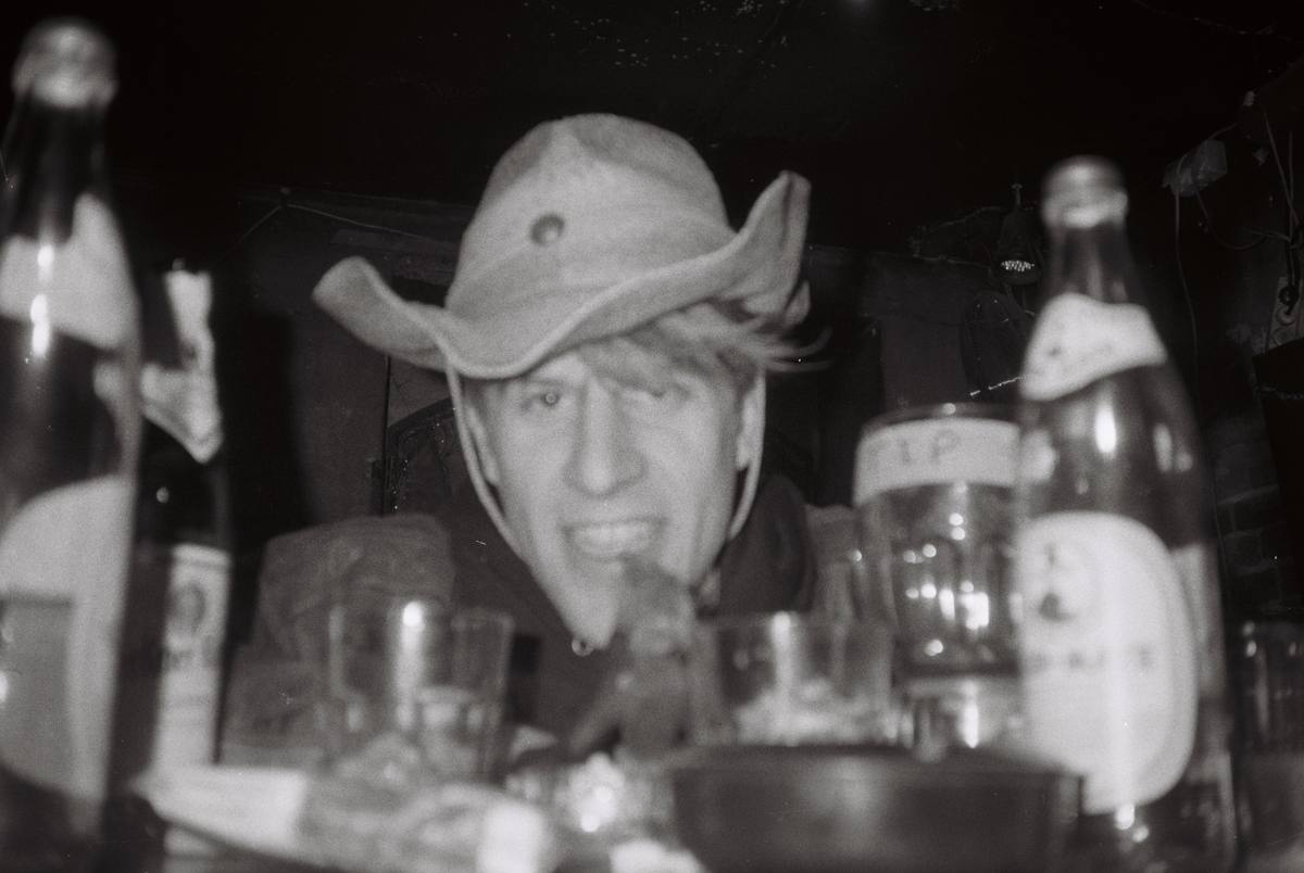 Black and white photo of Chris Pitsiokos wearing a hat at a table with bottles and glasses